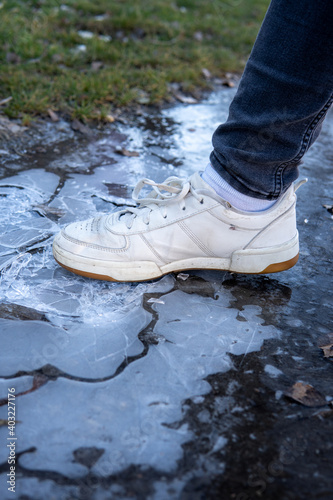 A woman in white sneakers breaking or crushing ice on a pathway in a park. Cracked ice under woman's feet on a cold, winter, day.