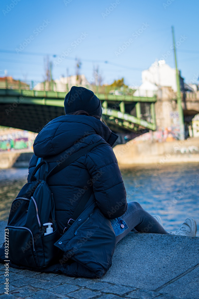 A girl sitting on the Danube river bank on a cold, windy day while wearing a black puffer coat, beanie and backpack. Green bridge and blue sky are shown in the background