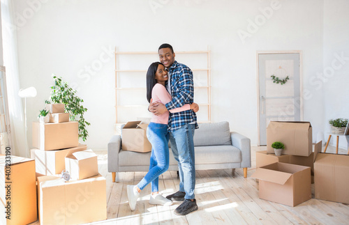 Romantic black newlywed couple hugging among cardboard boxes in their new apartment on relocation day, panorama