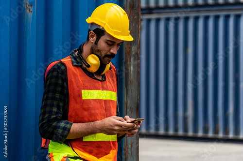 Caucasian worker in safety vest reflective with Safety helmet Typing a message on a cell phone in container pepot with Blue container box in background