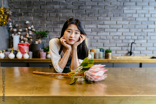 Open friendly asian woman lean on counter in modern interior of flower shop. Korean woman florist make floral decorations or compositions for sale.