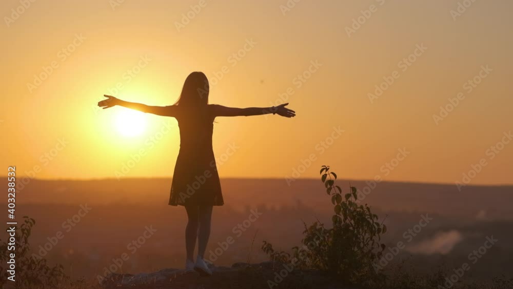 A young woman in summer dress jumping up with her raised hands outdoors enjoying view of bright yellow sunset. A young woman in summer dress jumping up with her raised hands outdoors enjoying view of bright yellow sunset.