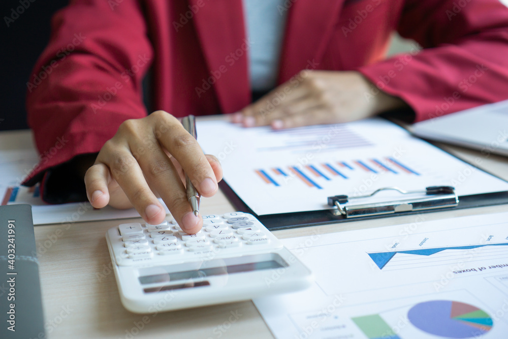 An Asian female finance worker is pressing on a calculator to calculate the company's earnings based on numbers and documents, bar graphs on her office desk, financial and profit report ideas. An Asian female finance worker is pressing on a calculator to calculate the company's earnings based on numbers and documents, bar graphs on her office desk, financial and profit report ideas.