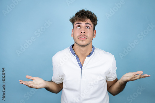 Young handsome man wearing a white t-shirt over blue background clueless and confused expression with arms and hands raised
