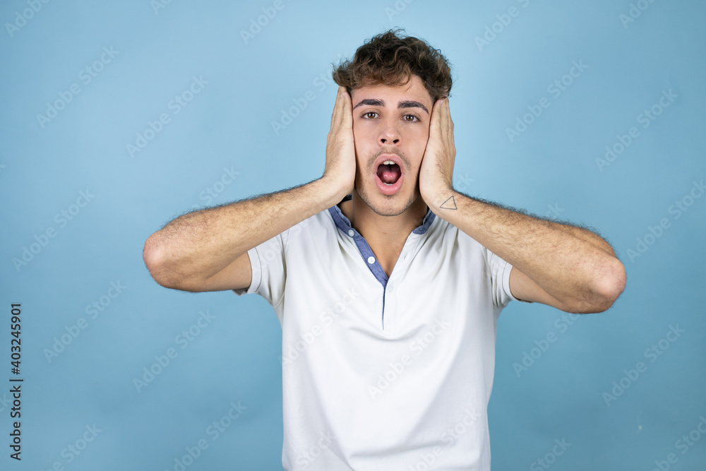 Young handsome man wearing a white t-shirt over blue background crazy and scared with hands on head, afraid and surprised of shock Young handsome man wearing a white t-shirt over blue background crazy and scared with hands on head, afraid and surprised of shock