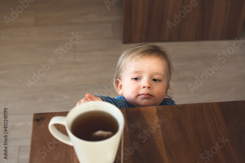 Little child try to grab cup of hot tea on the table. Attention hot content concept.