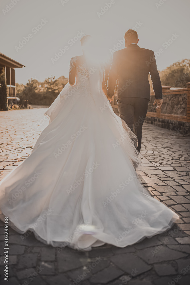 The wedding couple runs away into the sunset.The bride in a white long wedding dress is walking by the hand with the groom in a business classic suit. White sky dark gray brick floor on the background The wedding couple runs away into the sunset.The bride in a white long wedding dress is walking by the hand with the groom in a business classic suit. White sky dark gray brick floor on the background