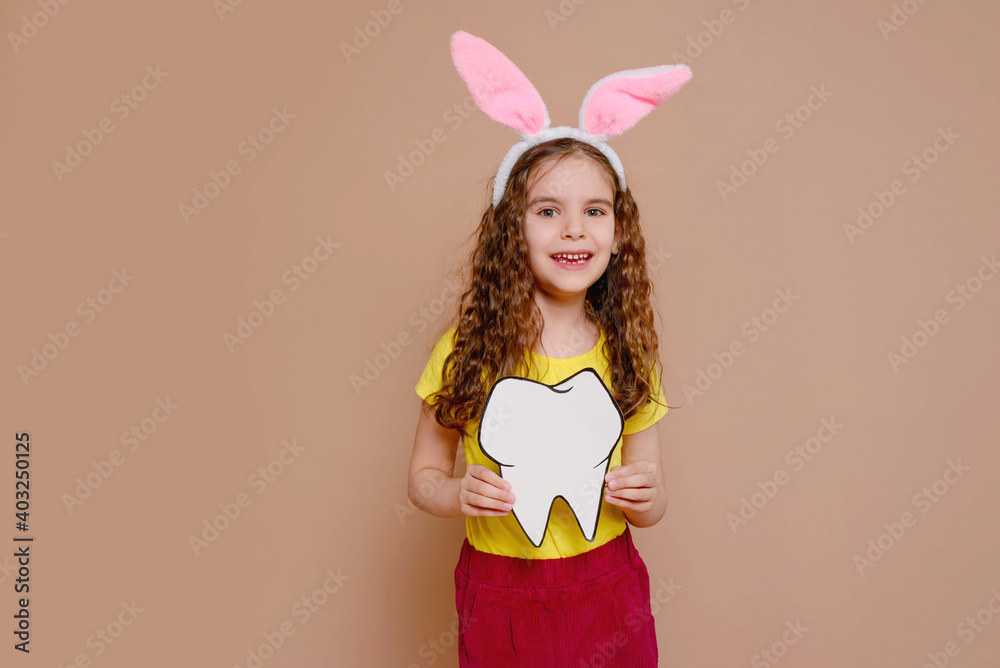 Cute blonde girl kid with rabbit ears holding a mock-up of a tooth. Traditions, discounts, medicine, the concept of the celebration. Cute blonde girl kid with rabbit ears holding a mock-up of a tooth. Traditions, discounts, medicine, the concept of the celebration.