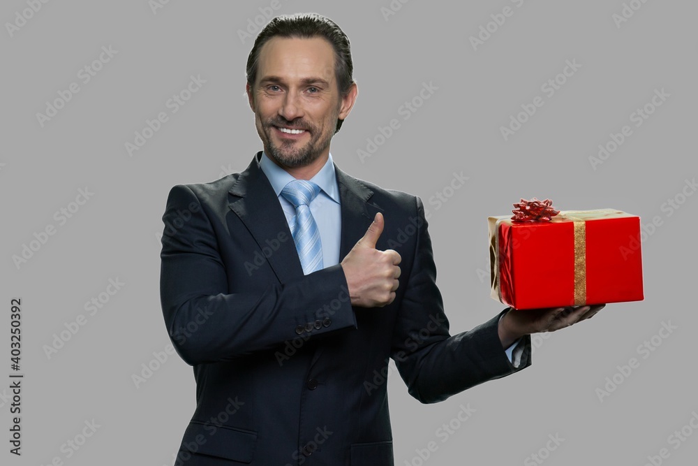 Happy businessman holding gift box and showing thumb up. Attractive caucasian man showing gift box on gray background. Happy businessman holding gift box and showing thumb up. Attractive caucasian man showing gift box on gray background.