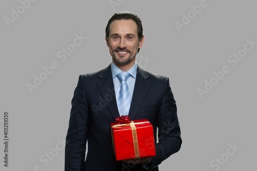 Handsome businessman holding gift box. Smiling caucasian man in business suit holding gift box on gray background. Holiday gift concept.