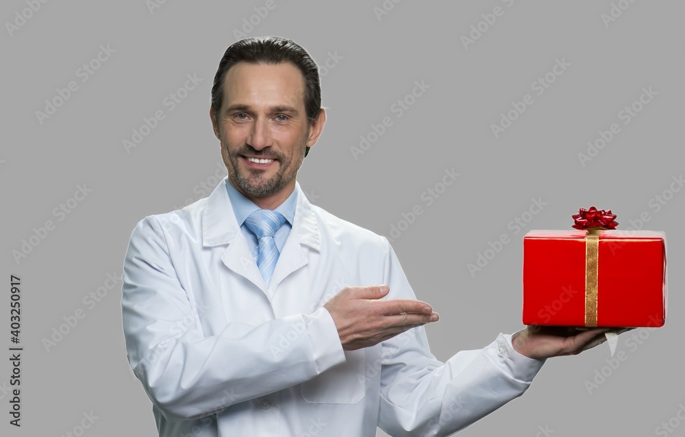 Portrait of smiling scientist showing gift box. Cheerful man in white coat offering red festive gift box against gray background. Portrait of smiling scientist showing gift box. Cheerful man in white coat offering red festive gift box against gray background.