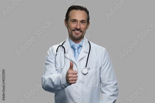 Portrait of smiling doctor showing thumb up gesture. Handsome male practitioner with stethoscope expressing success against gray background.