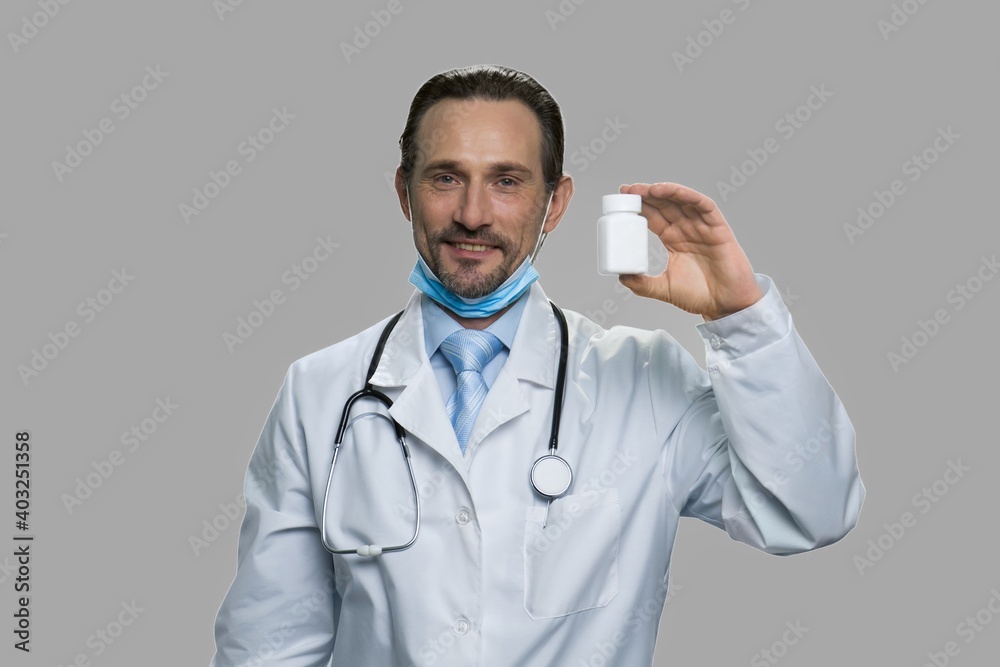 Professional male doctor showing bottle of pills. Attractive mature doctor with surgical mask and stethoscope against gray background. Professional male doctor showing bottle of pills. Attractive mature doctor with surgical mask and stethoscope against gray background.