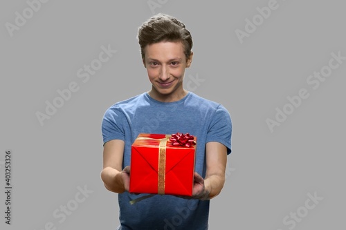 Handsome teen boy offering gift box to someone. Caucasian guy giving gift box to camera standing against gray background.