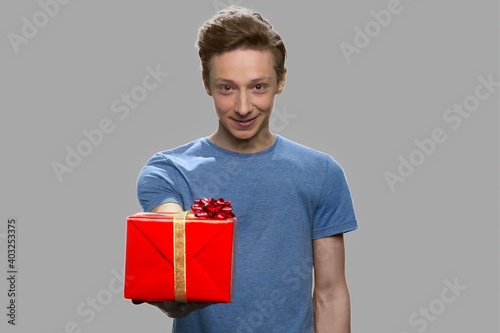 Teen guy giving gift box to camera. Handsome teenage boy offering present box on gray background.