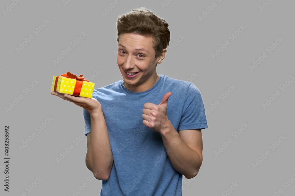 Funny teen boy holding gift box. Handsome cheerful teenage guy showing thumb up while holding gift box. Isolated on gray background. Funny teen boy holding gift box. Handsome cheerful teenage guy showing thumb up while holding gift box. Isolated on gray background.