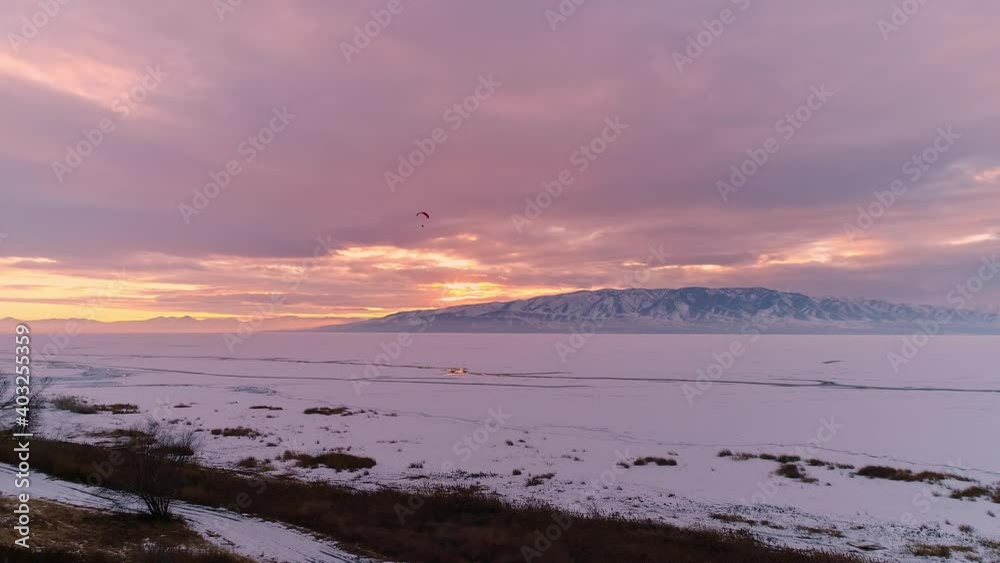 Flying towards frozen lake in winter landscape with paraglider flying against the sunset in Utah. Flying towards frozen lake in winter landscape with paraglider flying against the sunset in Utah.