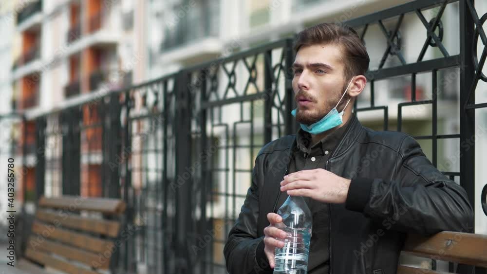 Young man sits on the bench, takes off medical protective mask and drinks mineral water from big blue plastic bottle Young man sits on the bench, takes off medical protective mask and drinks mineral water from big blue plastic bottle