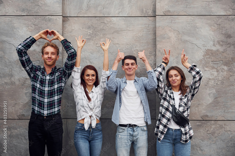 Making different gestures. Group of young positive friends in casual clothes standing together against grey wall