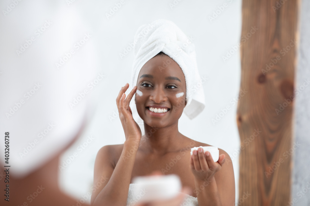 Smiling cute millennial african american lady with towel on head after shower applying cream on face Smiling cute millennial african american lady with towel on head after shower applying cream on face