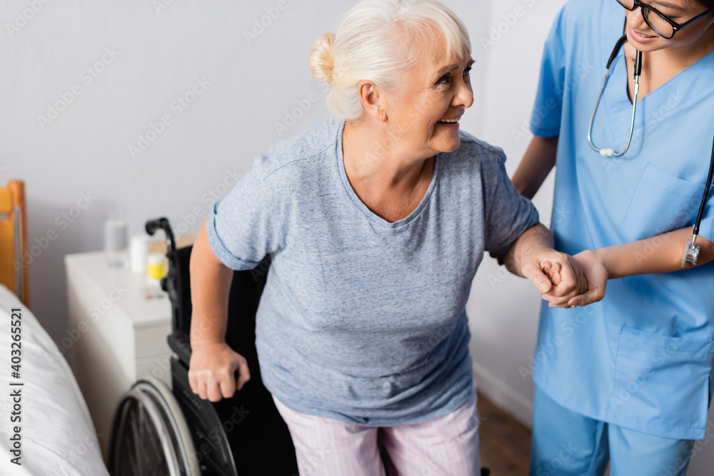 Obraz premium asian nurse supporting positive senior woman getting up from wheelchair