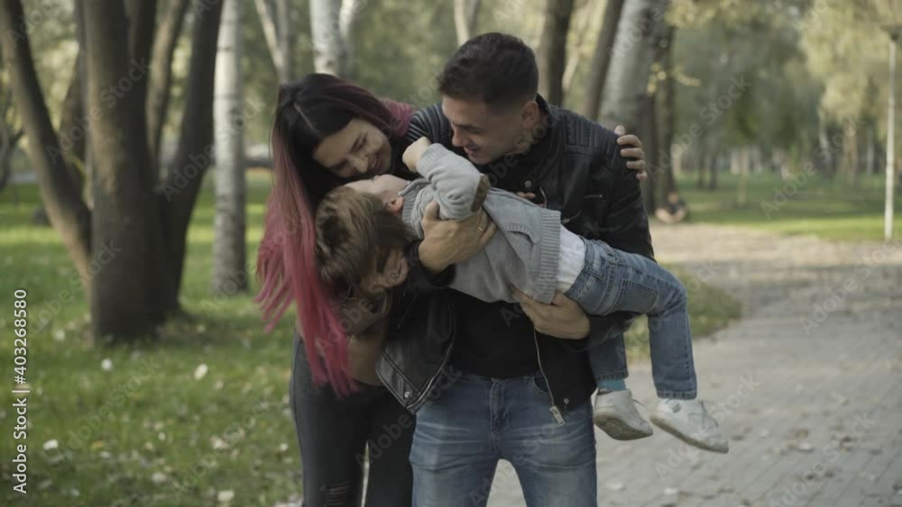 Positive handsome young father playing grimacing with little son as laughing mother hugging husband. Joyful Caucasian family having fun enjoying leisure outdoors on spring or summer day in park. Positive handsome young father playing grimacing with little son as laughing mother hugging husband. Joyful Caucasian family having fun enjoying leisure outdoors on spring or summer day in park.