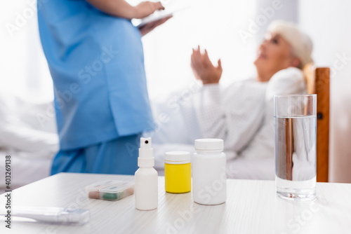 table with pills containers, throat spray and glass of water near nurse and patient on blurred foreground © LIGHTFIELD STUDIOS