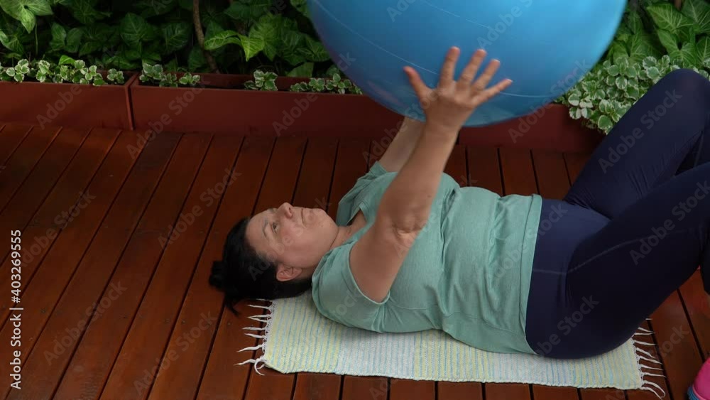 Mujer de mediana edad recostada en el piso haciendo ejercicio con pelota en el jardín de su casa