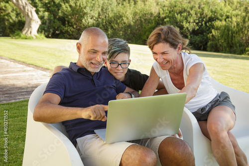 Padre , madre e figlio si divertono a guardare qualcosa di interessante nel computer portatile seduti in relax nel giardino di casa photo