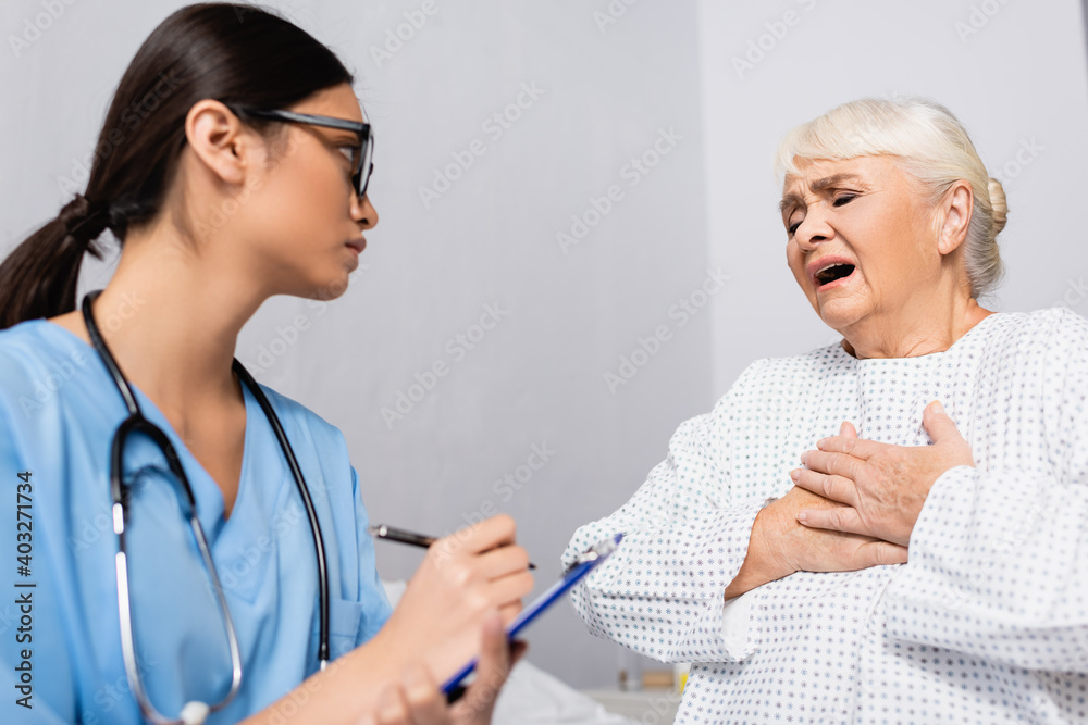Fototapeta premium aged woman touching chest while suffering from asthma attack near asian nurse writing on clipboard, blurred foreground