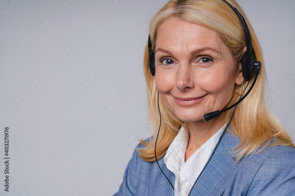 Aged beautiful call center worker in formal attire with headphones isolated over grey background Aged beautiful call center worker in formal attire with headphones isolated over grey background