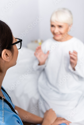 young nurse listening to cheerful aged woman gesturing while talking on blurred background