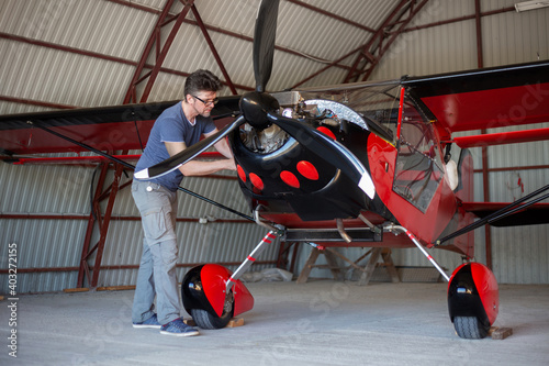 Repairman looking over engine in small aircraft from below. Small red airplane in hangar. Repaiper holding instruments in the hand near the plane engine. Small aviation.