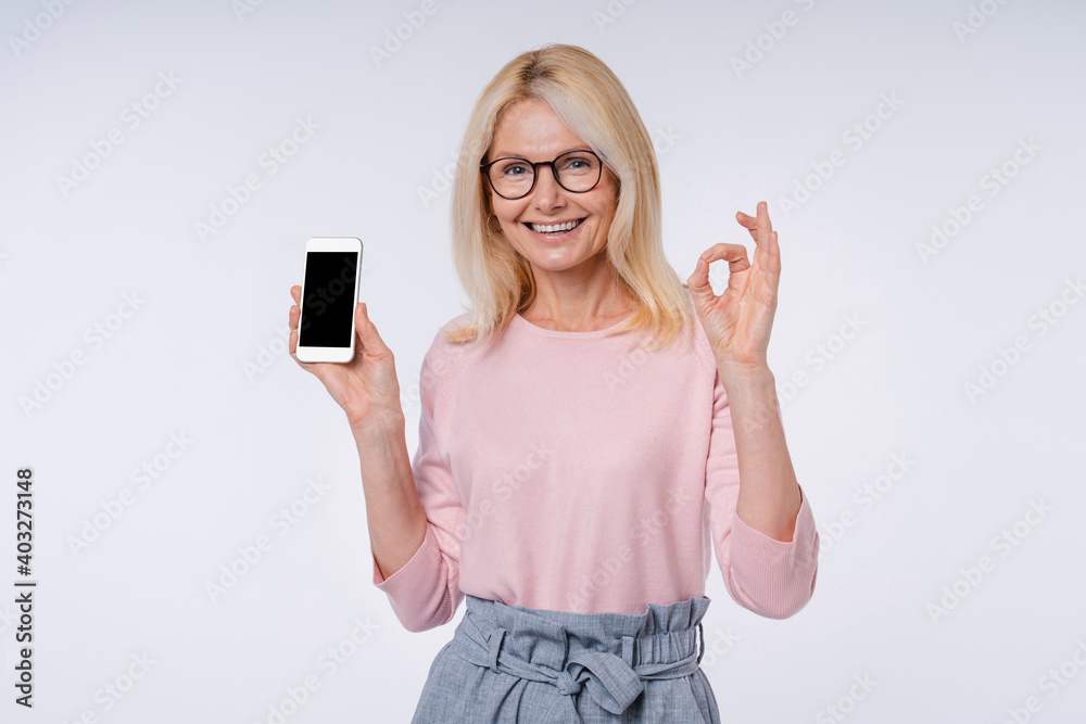 Smart elderly woman holding mobile phone and showing okay sign isolated over grey background Smart elderly woman holding mobile phone and showing okay sign isolated over grey background