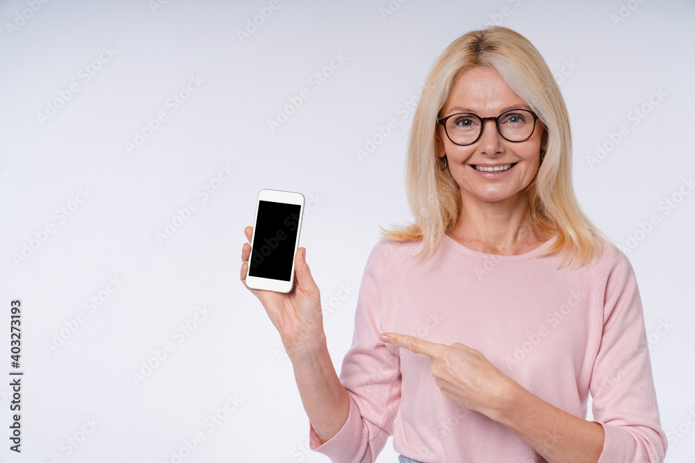 Attractive mature european woman pointing at smart phone in casual attire isolated over grey background Attractive mature european woman pointing at smart phone in casual attire isolated over grey background