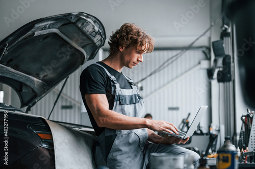 With laptop. Adult man in grey colored uniform works in the automobile salon