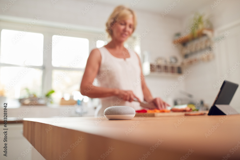 Retired Woman Making Meal In Kitchen With Smart Speaker In Foreground Retired Woman Making Meal In Kitchen With Smart Speaker In Foreground