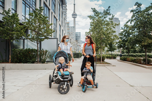 Two Caucasian moms with strollers and kids walking together in Toronto city Canada. Women in face masks with children outdoor. Friends talking on street keeping social distance. A new normal.