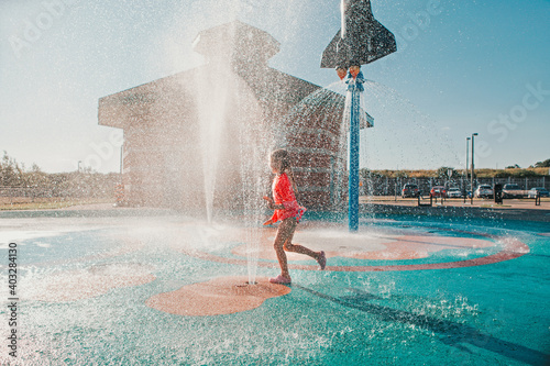 Cute adorable Caucasian funny girl playing on splash pad playground on summer day. Happy child having fun in water. Seasonal water sport recreational activity for kids outdoors. photo