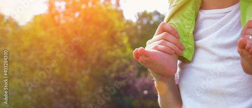 A man with a child on his shoulders. Close-up of barefoot little leg and hand of father on the background of nature trees and sun rays. Banner. concept of a family outdoor recreation. Soft focus.