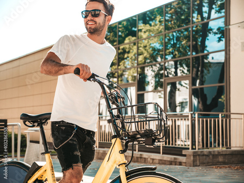 Portrait of handsome smiling stylish hipster lambersexual model.Man dressed in white T-shirt. Fashion male riding a bike on the street background in sunglasses © halayalex