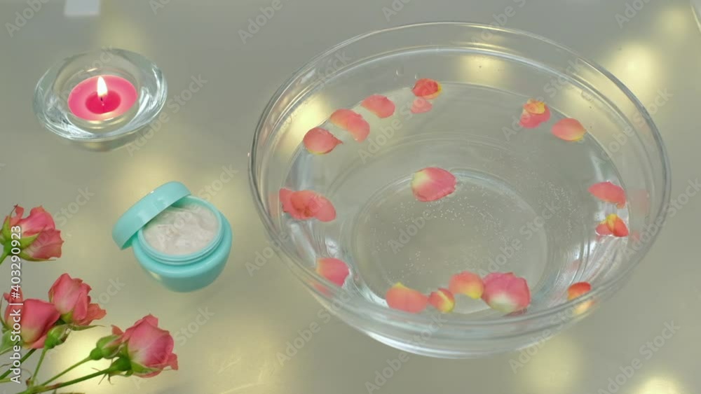 A rose petal falls into the water and forms a wave inside a glass bowl, next to the bowl are hand cream, a bouquet of small roses and a candle is burning. A rose petal falls into the water and forms a wave inside a glass bowl, next to the bowl are hand cream, a bouquet of small roses and a candle is burning.