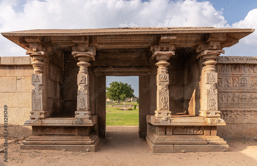Fototapeta premium Hampi, Karnataka, India - November 4, 2013: Hazara Rama Temple. Beige stone entrance hall with flat roof and fresco decorated pillars is set in outside wall, showing green foliage and blue cloudscape.