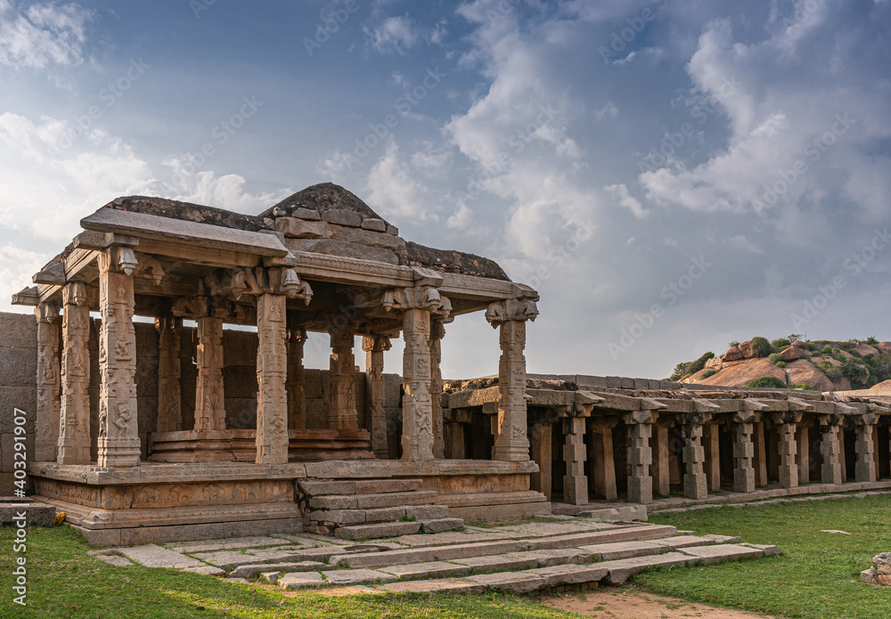 Hampi, Karnataka, India - November 4, 2013: Hazara Rama Temple. Ruinous brown stone buildings along outside walls with rows of columns under blue cloudscape with grass in front.