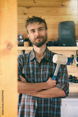 Portrait of smiling brutal bearded worker man stands in workshop with mallet, construction carpentry tools in the background. DIY concept. Vertical.