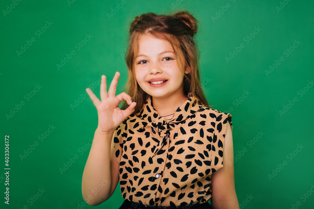 Teenage girl in colorful clothes on a green background with a charming smile. The child shows the gesture ok. Teenage girl in colorful clothes on a green background with a charming smile. The child shows the gesture ok.