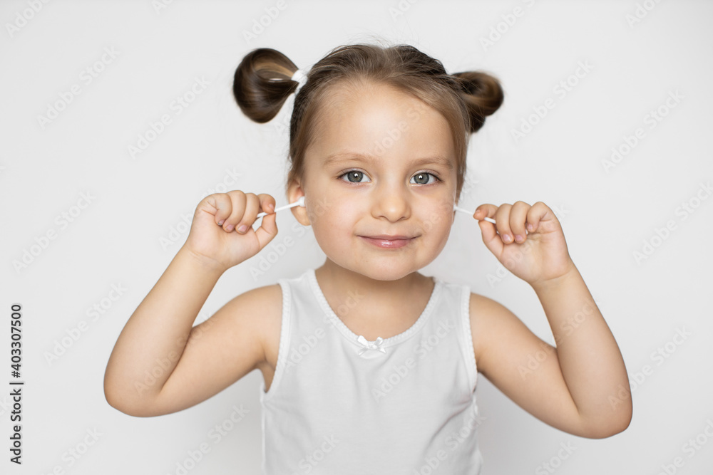 Closeup photo of charming little baby girl, wearing white tank top, cleaning her ears with cotton swabs, posing over the white studio background. Ear hygiene concept Closeup photo of charming little baby girl, wearing white tank top, cleaning her ears with cotton swabs, posing over the white studio background. Ear hygiene concept