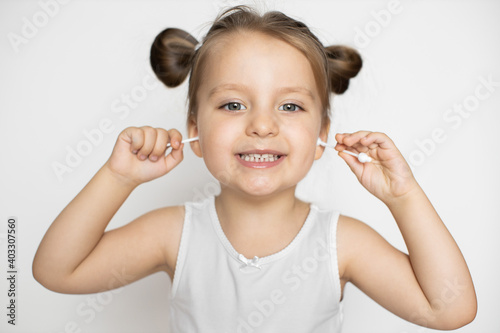 Close up portrait of happy laughing three years old kid girl, posing to camera with teeth smile and cleaning her little ears with cotton swabs. Skincare and ear hygiene