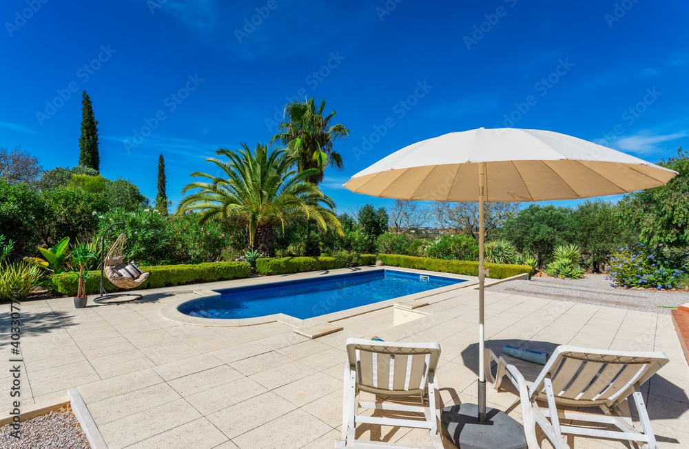 Pool with poolside umbrella and sun loungers. Clear blue sky and beautiful trees in the background on a nice summer day.