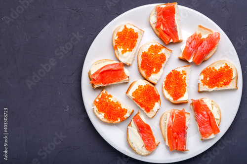 Bruschettes with butter and red caviar and salmon on a white plate on a gray concrete background copy space.
